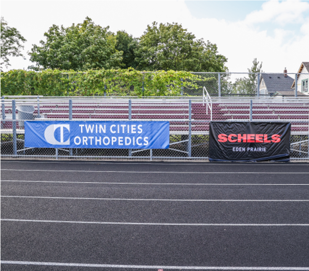 Photo of South end of Al Gowans stadium bleachers showing the fence by the track with spnsors banners: Twin Cities Orthopedics and Scheels