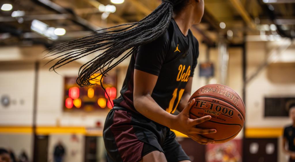image of Minneapolis Roosevelt High School girls basketball player dribbling the ball up the court after a rebound. Behind her, in soft focus, is the wall cushion with “ROOSEVELT” in gold block letters printed on it.