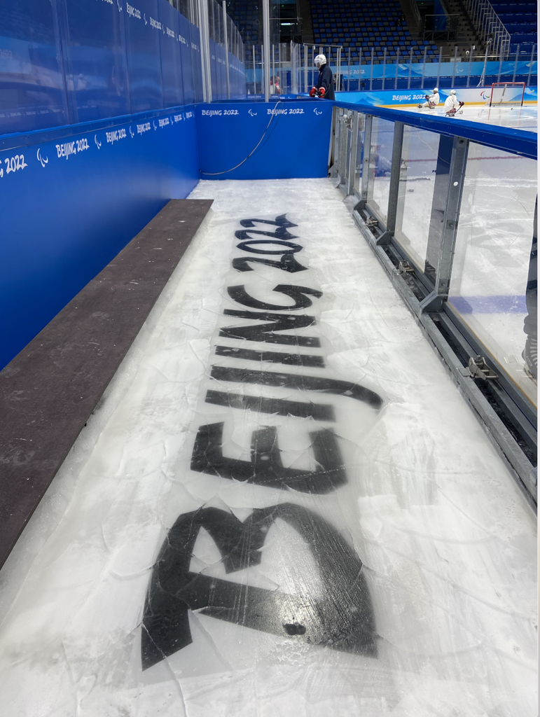 A sled hockey bench with real ice and transparent boards. The ice has Beijing 2022 printed on it in black.